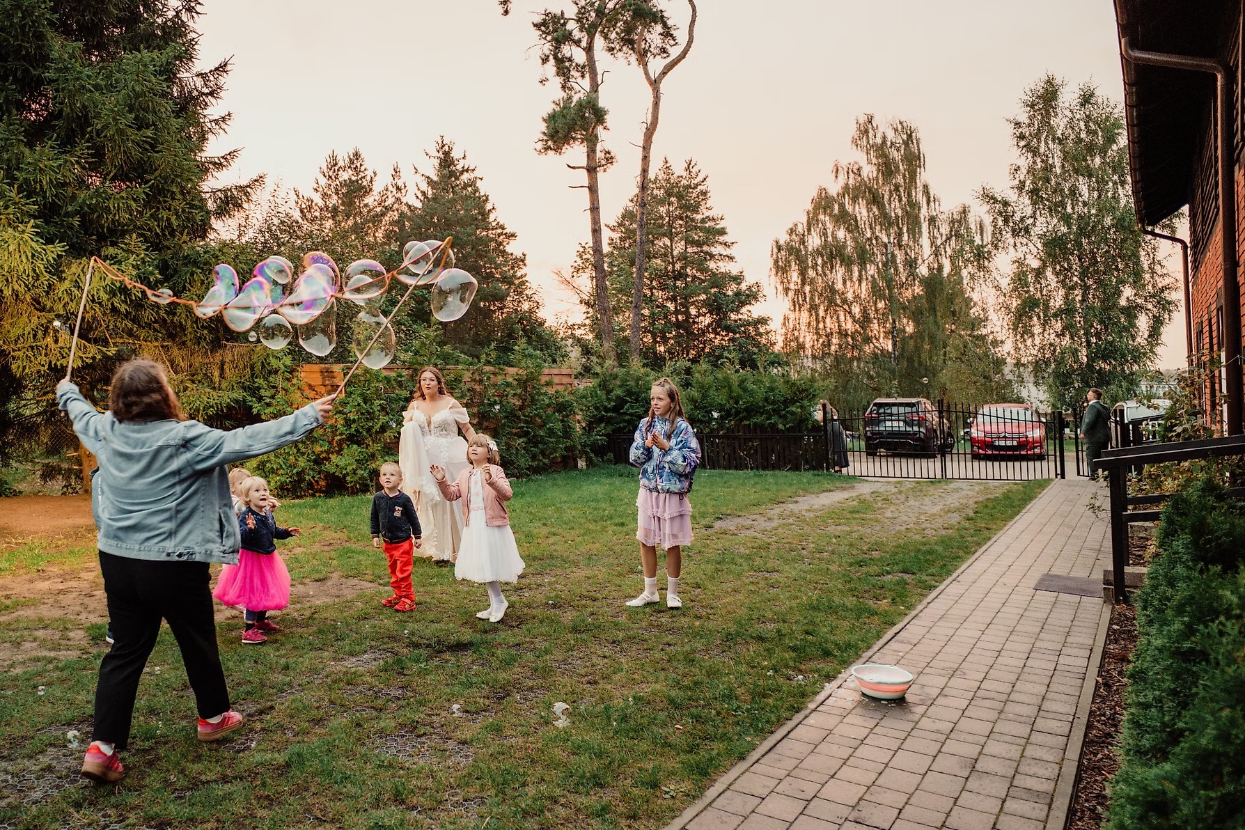 Animator puszcza bańki mydlane dla dzieci na przyjęciu w ogrodzie. W tle kobieta w białej sukni i goście. Ujęcie z perspektywy osoby puszczającej bańki.