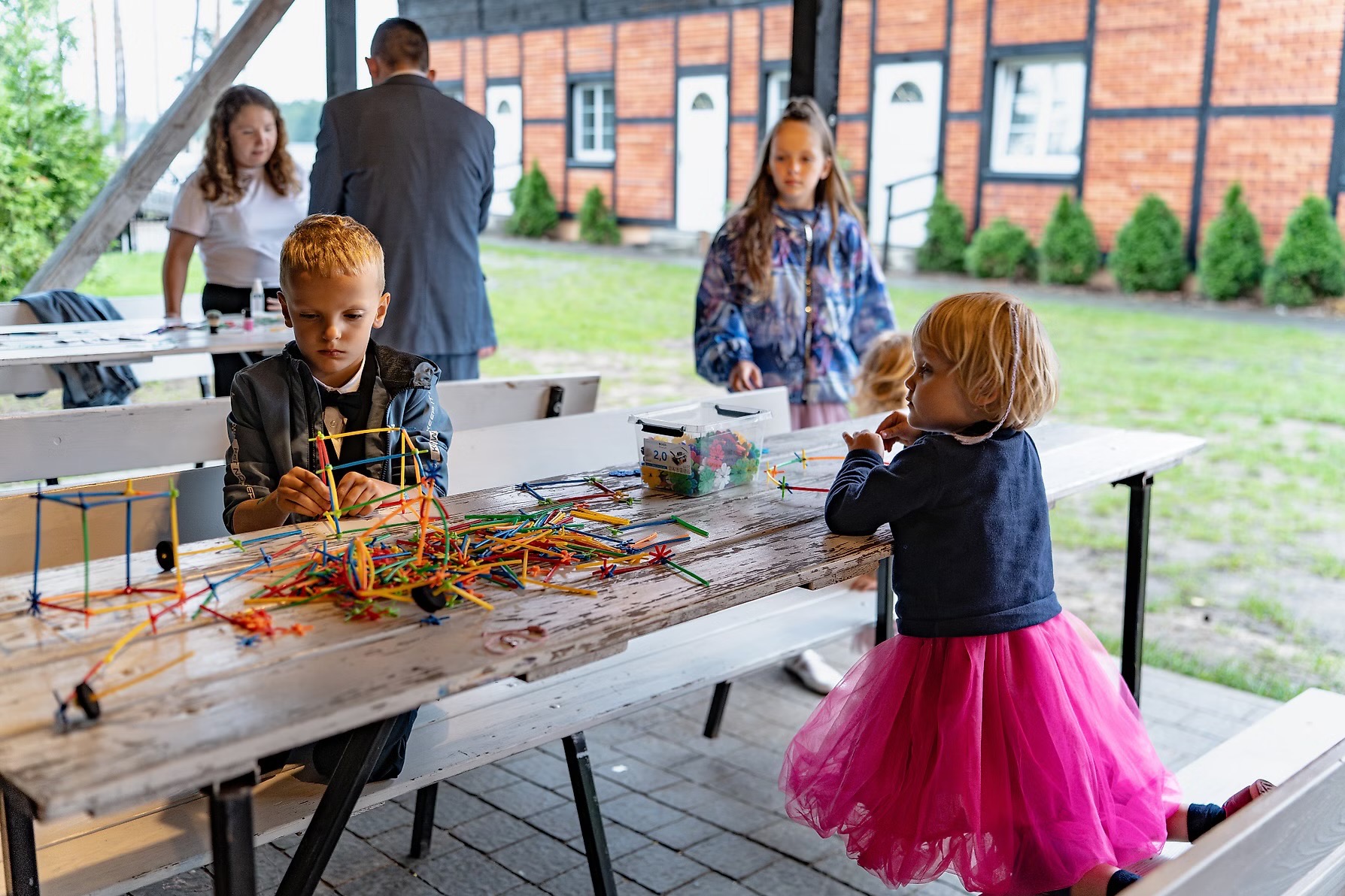 Dzieci przy drewnianym stole budują konstrukcje z kolorowych patyczków. W tle budynek z czerwonej cegły i zielone krzewy. Dziewczynka w różowej spódniczce.