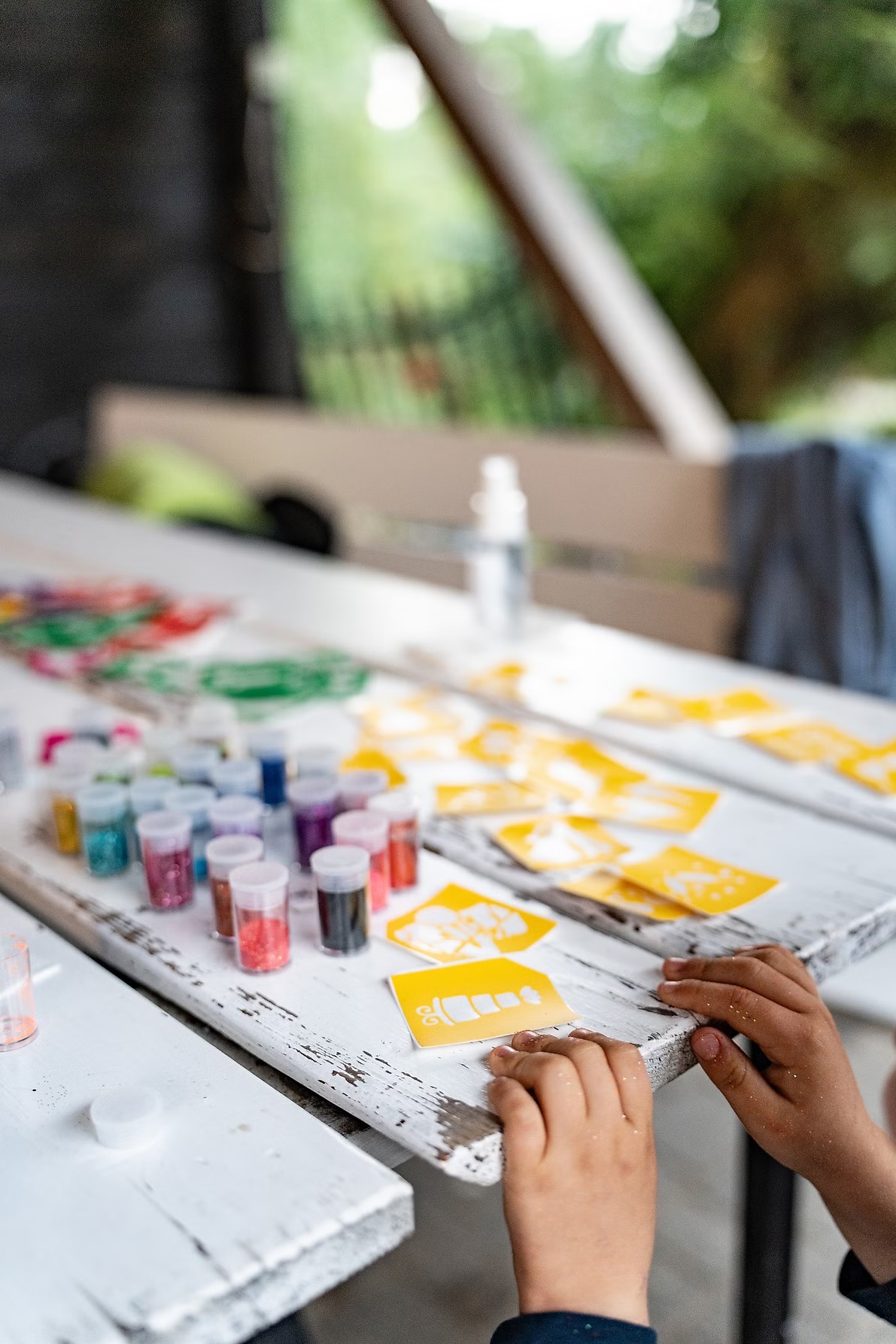 Dziecięce dłonie przy białym stole z brokatem, farbami i szablonami. Przygotowania do kreatywnych zajęć plastycznych na świeżym powietrzu.