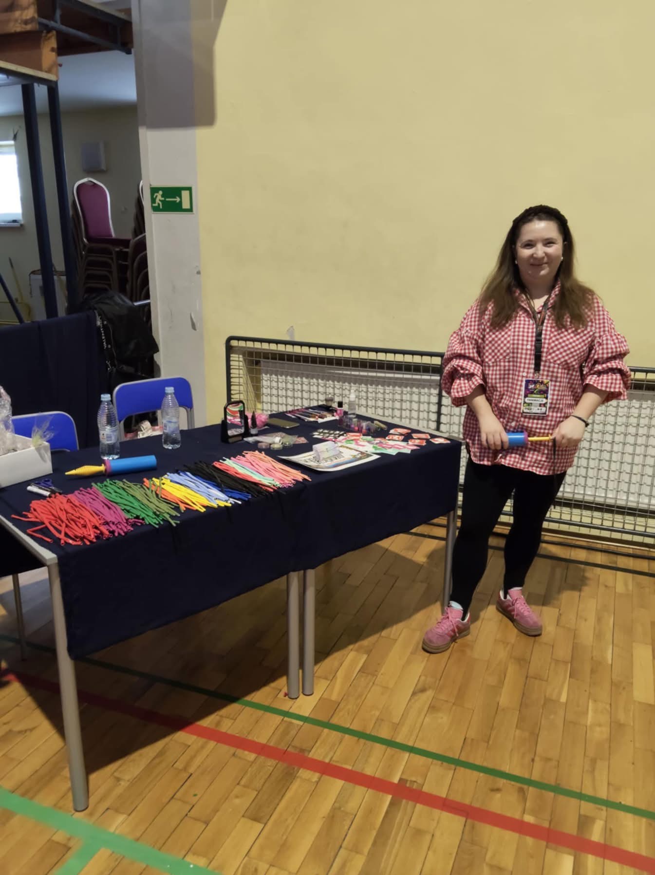 Animator prezentuje kolorowe balony i akcesoria na stole, przygotowując się do animacji. W tle widoczna ściana i podłoga sali gimnastycznej.