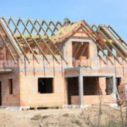 Niedokończony dom murowany z widoczną konstrukcją dachu i częściowo wymurowanymi ścianami z bloczków ceramicznych, na tle błękitnego nieba.