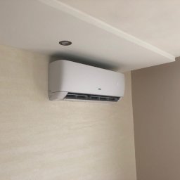 COOLBIX Tomasz Kulbicki - Biała klimatyzacja ścienna z widocznym panelem sterowania, zamontowana w jasnym pomieszczeniu z beżowymi ścianami i oświetleniem punktowym w suficie.