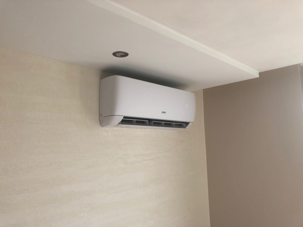 Biała klimatyzacja ścienna z widocznym panelem sterowania, zamontowana w jasnym pomieszczeniu z beżowymi ścianami i oświetleniem punktowym w suficie.