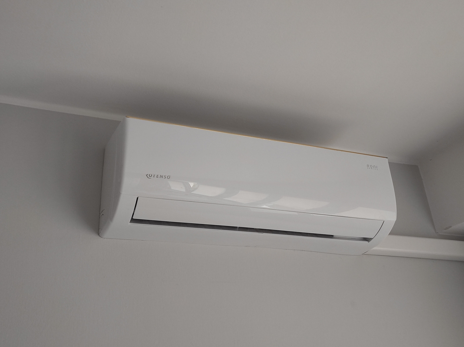 Biała klimatyzacja Rulenso Roni Series zamontowana na szarej ścianie, widok z dołu, delikatne oświetlenie, minimalistyczny design.