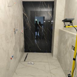 Marbax - Wykończona łazienka z czarną kabiną prysznicową i marmurowymi płytkami na ścianach i podłodze. Widoczny odpływ liniowy i narzędzia glazurnicze.