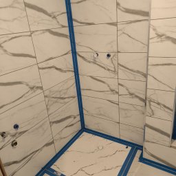 Marbax - Wykończona kabina prysznicowa z białymi płytkami imitującymi marmur, zabezpieczona niebieską taśmą malarską. Widoczne otwory na armaturę.