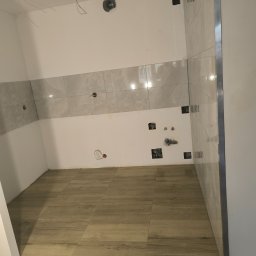 Marbax - Wnętrze łazienki w trakcie prac wykończeniowych: ułożone płytki podłogowe imitujące drewno i częściowo położone szare płytki ścienne z widocznymi puszkami elektrycznymi.
