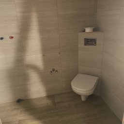 Marbax - Wykończona łazienka z beżowymi płytkami imitującymi drewno, wisząca toaleta z zabudowaną spłuczką. Widoczne przyłącza wodne i kanalizacyjne. W tle fragment drabiny.