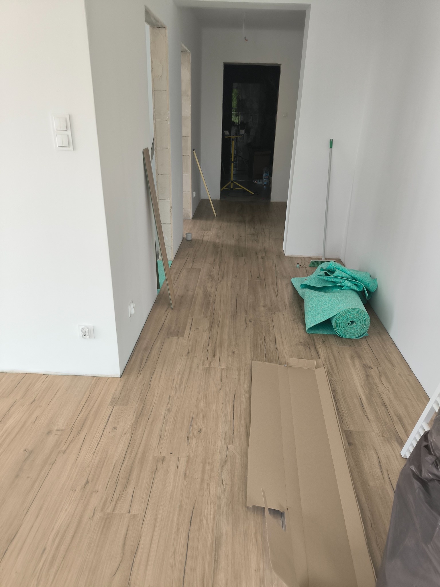 Jasne panele podłogowe w korytarzu po remoncie. Widoczne materiały budowlane i narzędzia. Świeżo pomalowane ściany na biało. Otwory drzwiowe bez ościeżnic.