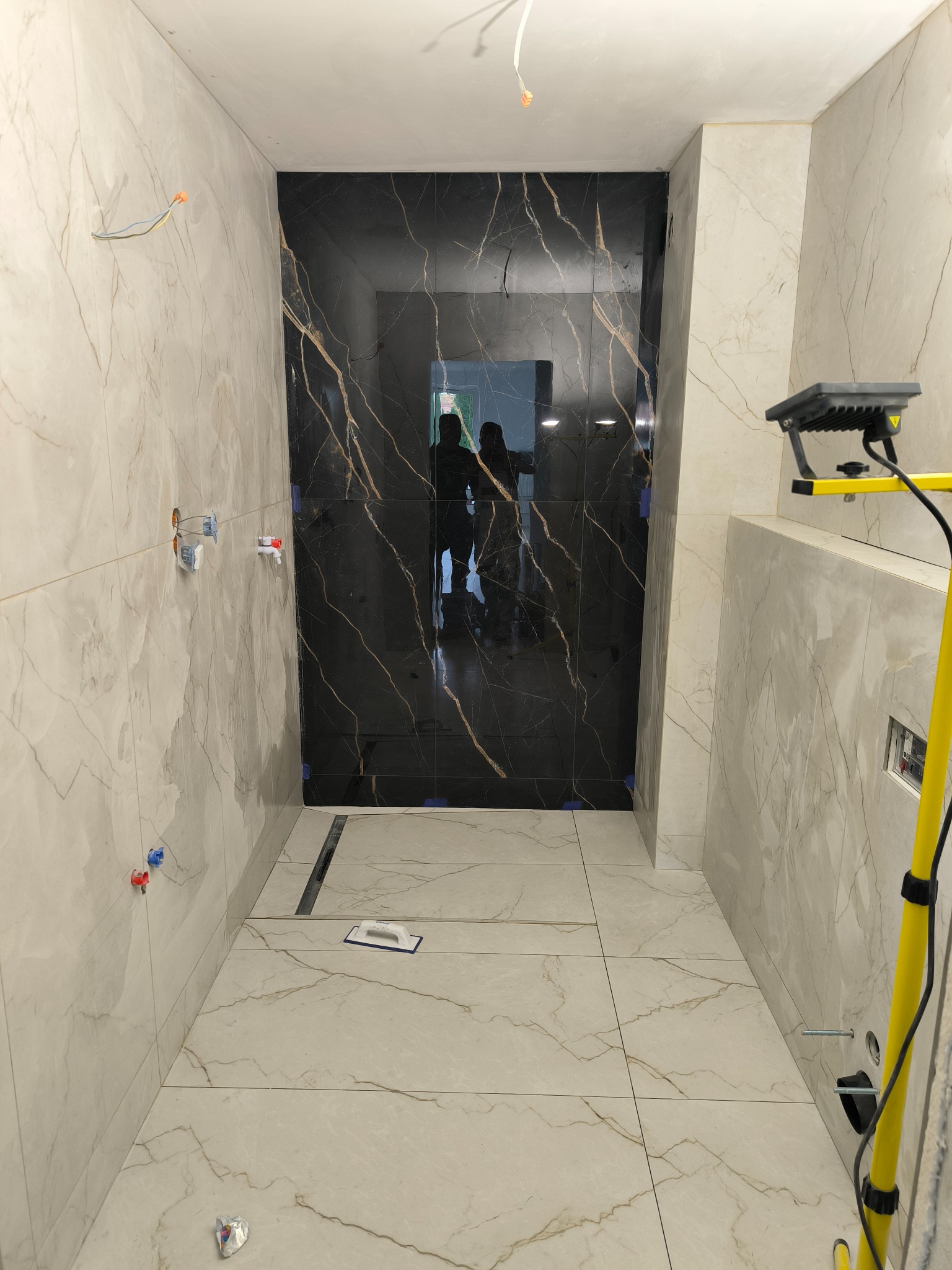 Wykończona łazienka z czarną kabiną prysznicową i marmurowymi płytkami na ścianach i podłodze. Widoczny odpływ liniowy i narzędzia glazurnicze.