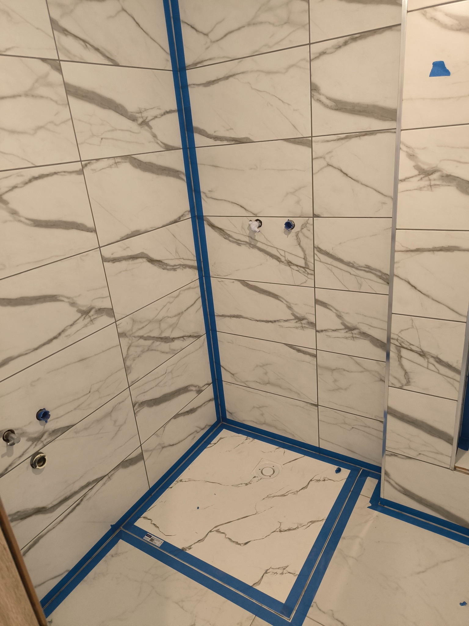 Wykończona kabina prysznicowa z białymi płytkami imitującymi marmur, zabezpieczona niebieską taśmą malarską. Widoczne otwory na armaturę.