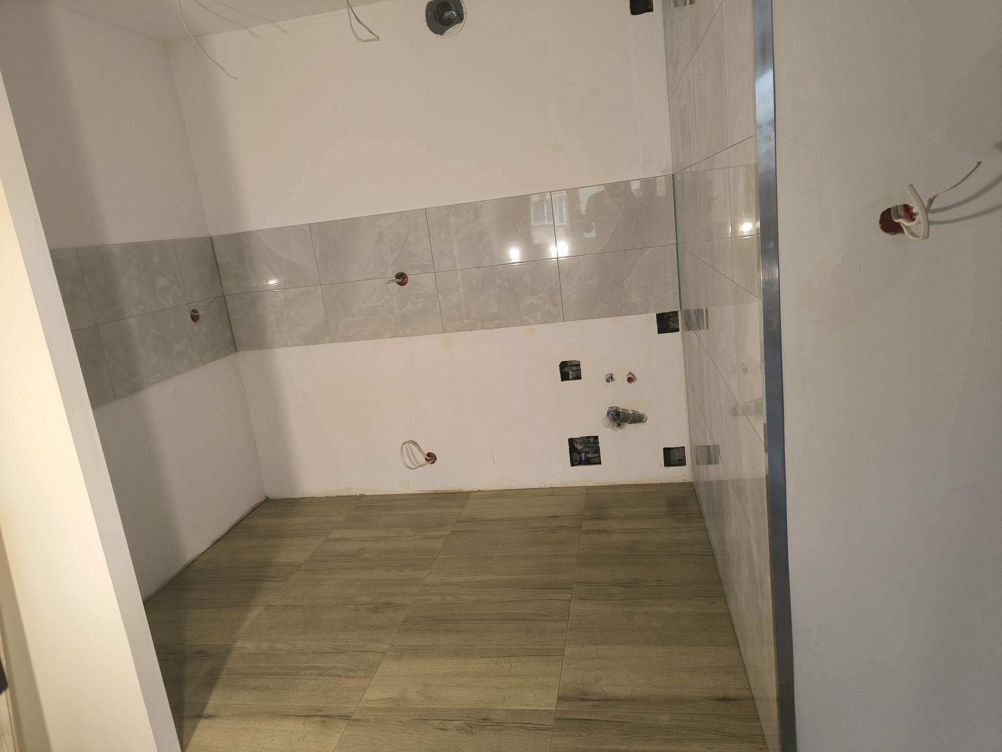 Wnętrze łazienki w trakcie prac wykończeniowych: ułożone płytki podłogowe imitujące drewno i częściowo położone szare płytki ścienne z widocznymi puszkami elektrycznymi.