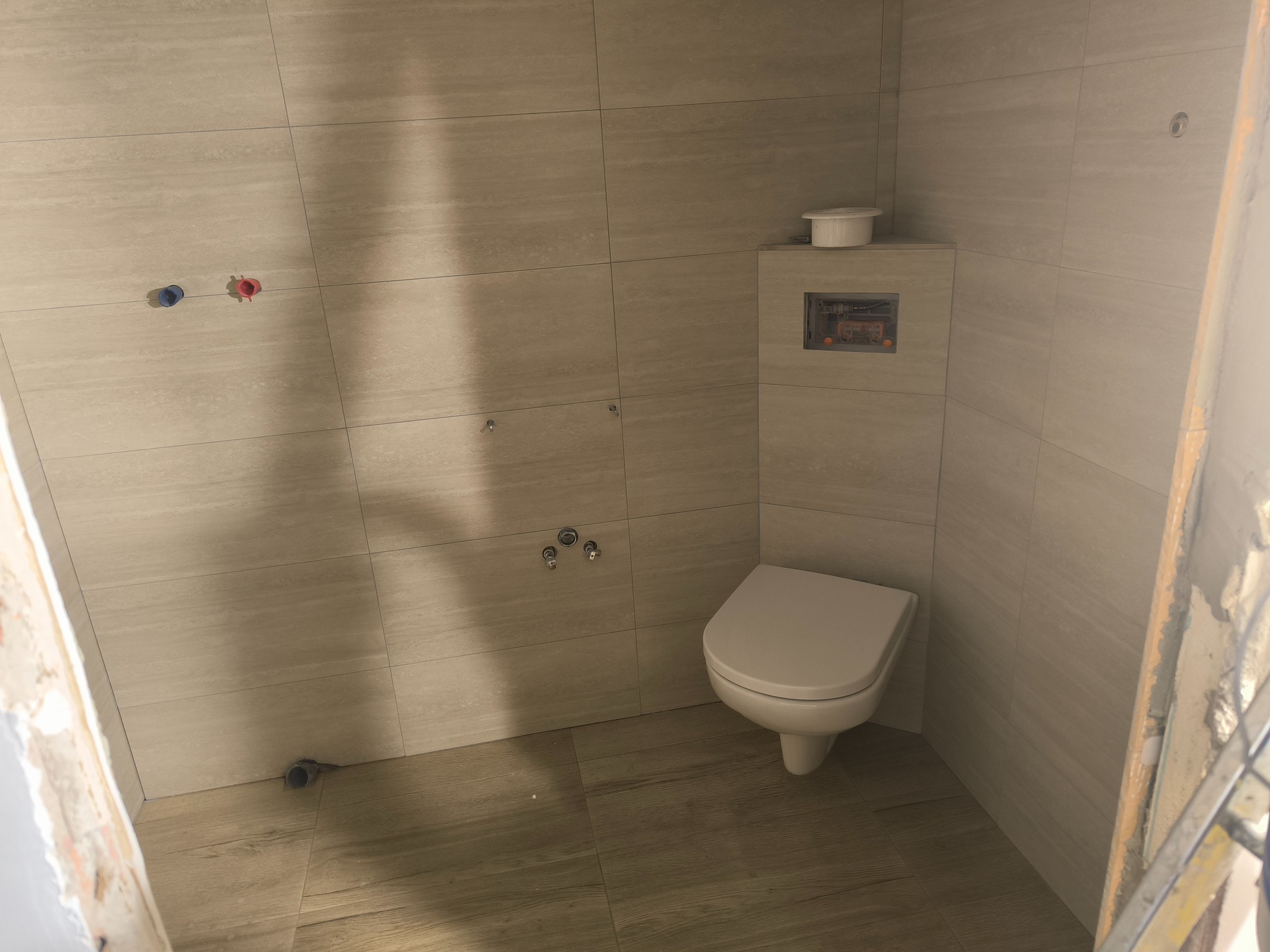 Wykończona łazienka z beżowymi płytkami imitującymi drewno, wisząca toaleta z zabudowaną spłuczką. Widoczne przyłącza wodne i kanalizacyjne. W tle fragment drabiny.