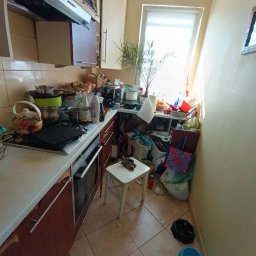 Sylwia Chwałowska - Zaniedbana kuchnia z widocznym bałaganem na blacie, zlewie i podłodze. Naczynia, garnki, środki czystości i inne przedmioty tworzą nieporządek. Widoczne miski dla zwierząt.