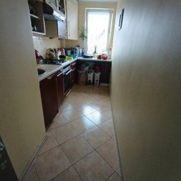 Sylwia Chwałowska - Ustawna kuchnia w bloku po sprzątaniu: brązowe szafki, beżowe ściany, okno z rośliną. Widok na czystą podłogę z płytek i uporządkowany blat.