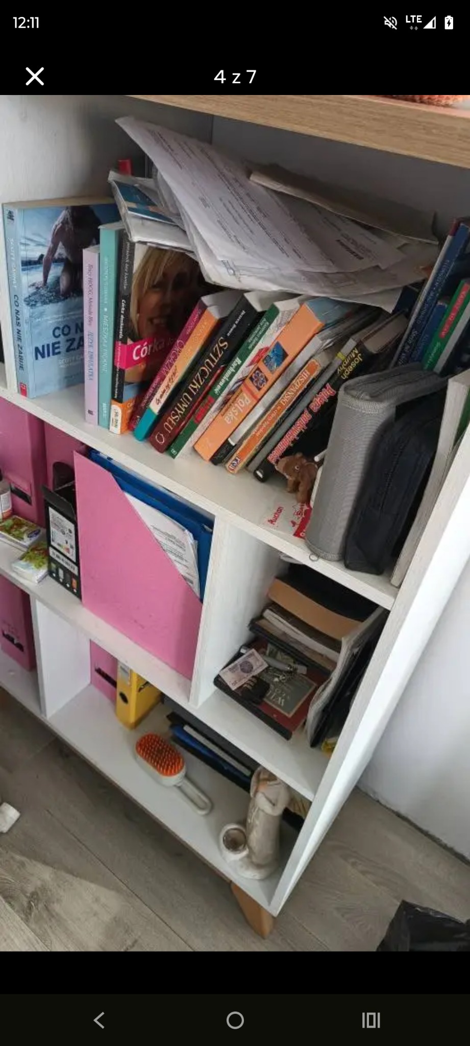 Półka regału z książkami, dokumentami i drobnymi przedmiotami w nieładzie. Widoczne segregatory, książki o różnej tematyce i przedmioty codziennego użytku.