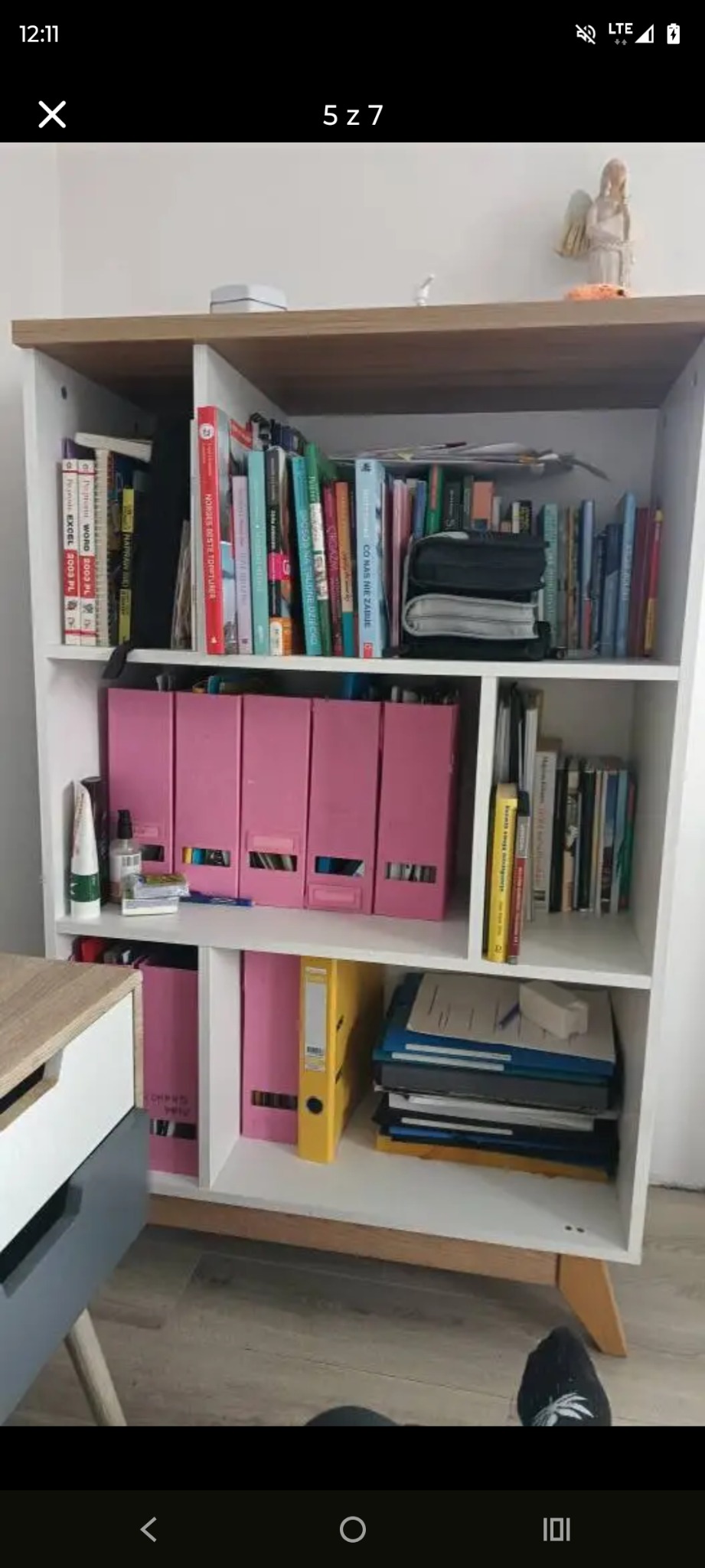 Półka z książkami, segregatorami i dekoracjami. Widoczne książki, segregatory w odcieniach różu i żółci, oraz drobne przedmioty dekoracyjne na górnej półce.