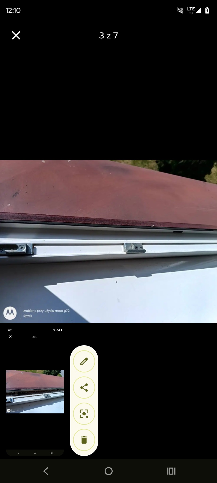 Brudne okno dachowe z widocznymi zaciekami i osadami, uchwycone z bliska pod kątem. Widoczne elementy mocujące i zarys zieleni w tle.