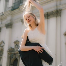 trzaska.fotografia - Uśmiechnięta panna młoda w koronkowej sukni, trzymająca bukiet nad głową, niesiona przez pana młodego na tle jasnej fasady budynku.