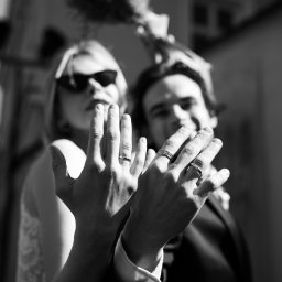 trzaska.fotografia - Czarno-białe zbliżenie dłoni pary z obrączkami ślubnymi, panna młoda w okularach przeciwsłonecznych, bukiet w tle, podkreślające uroczysty charakter.