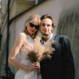 trzaska.fotografia - Para młoda w objęciach, panna młoda w koronkowej sukni trzyma bukiet z traw, pan młody w garniturze z wzorzystą apaszką, miejska sceneria w tle.