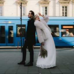 trzaska.fotografia - Para młoda w miejskiej scenerii: panna młoda w futrze opiera się o pana młodego na tle przejeżdżającego, rozmazanego tramwaju. Nietypowe ujęcie ślubne.
