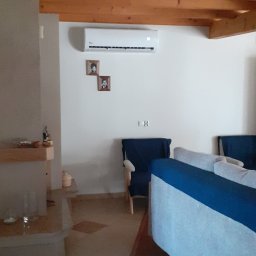 SniMer Piotr Gajos - Biały klimatyzator na ścianie w salonie z drewnianym sufitem. Pod spodem dwie małe ramki ze zdjęciami. W tle niebieskie fotele i fragment sofy.