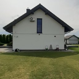 SniMer Piotr Gajos - Biały dom z ciemnym dachem i zamontowaną klimatyzacją na ścianie. Widok z trawnika, w tle budynek gospodarczy i drzewa.