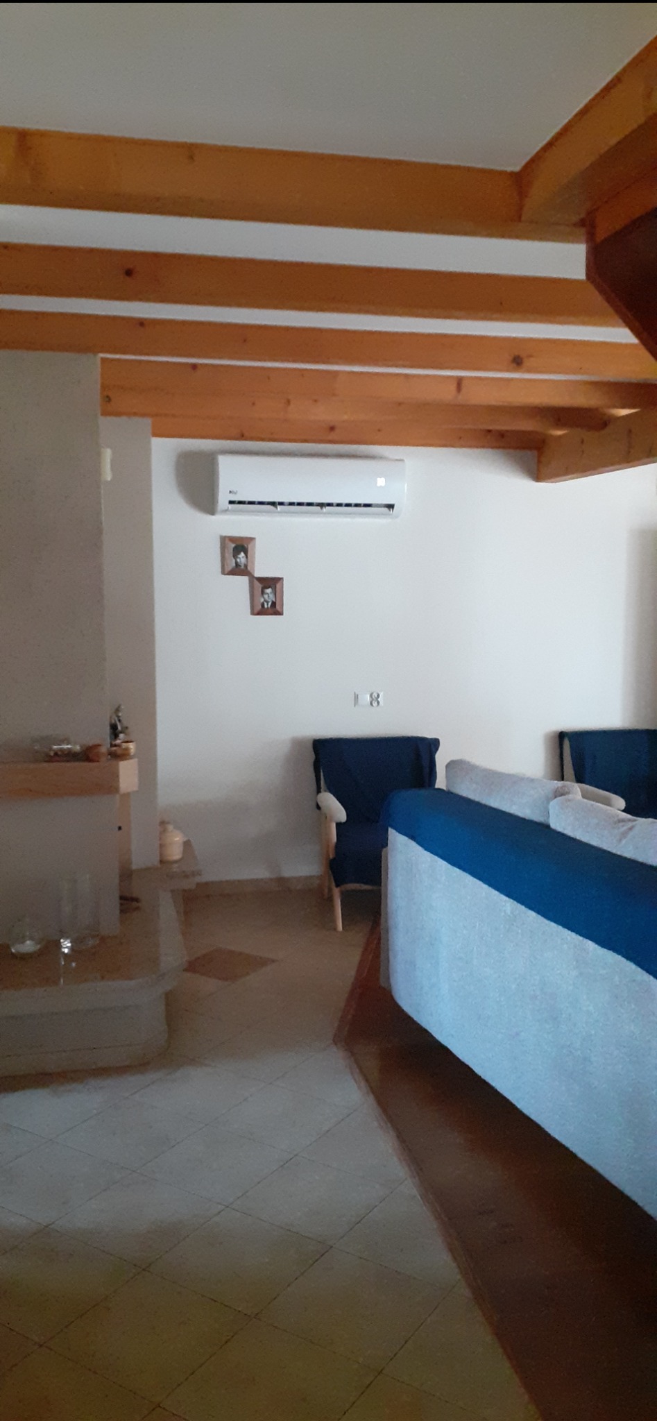 Biały klimatyzator na ścianie w salonie z drewnianym sufitem. Pod spodem dwie małe ramki ze zdjęciami. W tle niebieskie fotele i fragment sofy.