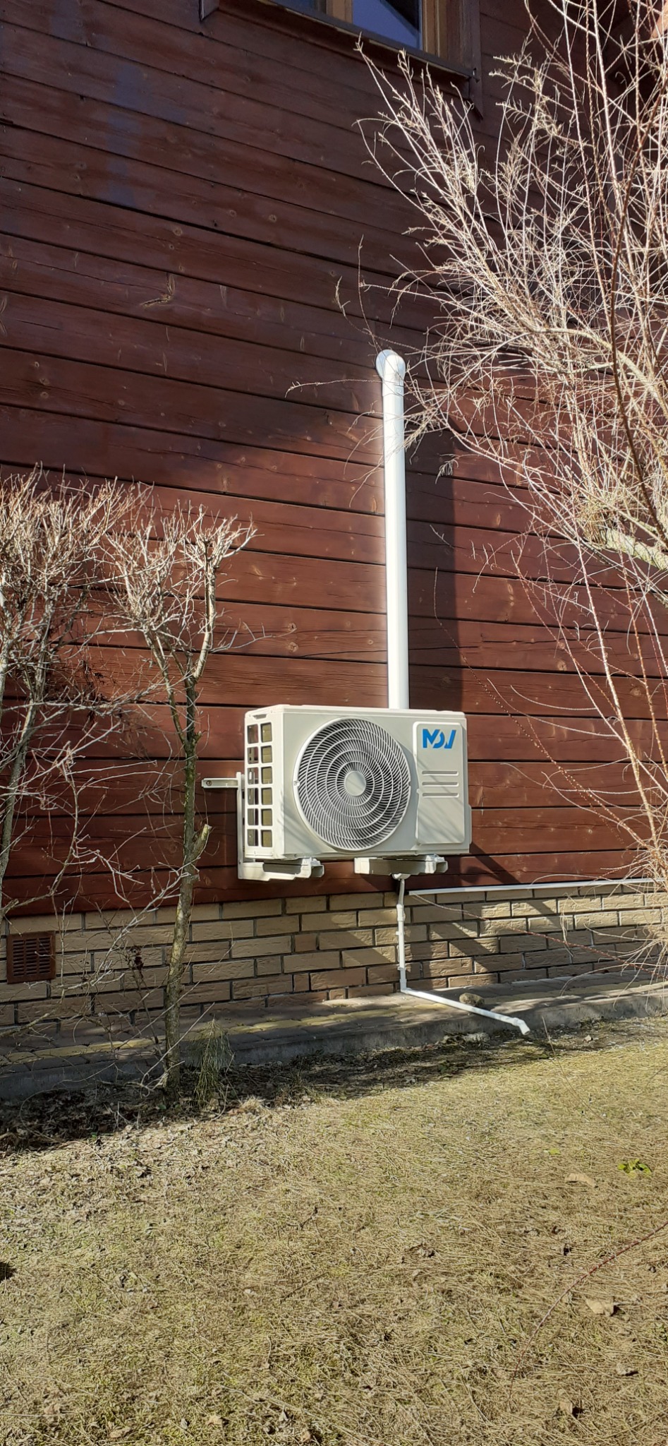 Klimatyzator zamontowany na brązowej, drewnianej ścianie domu, z widocznymi rurami odprowadzającymi. Obok krzew z gołymi gałęziami. Ujęcie w słoneczny dzień.