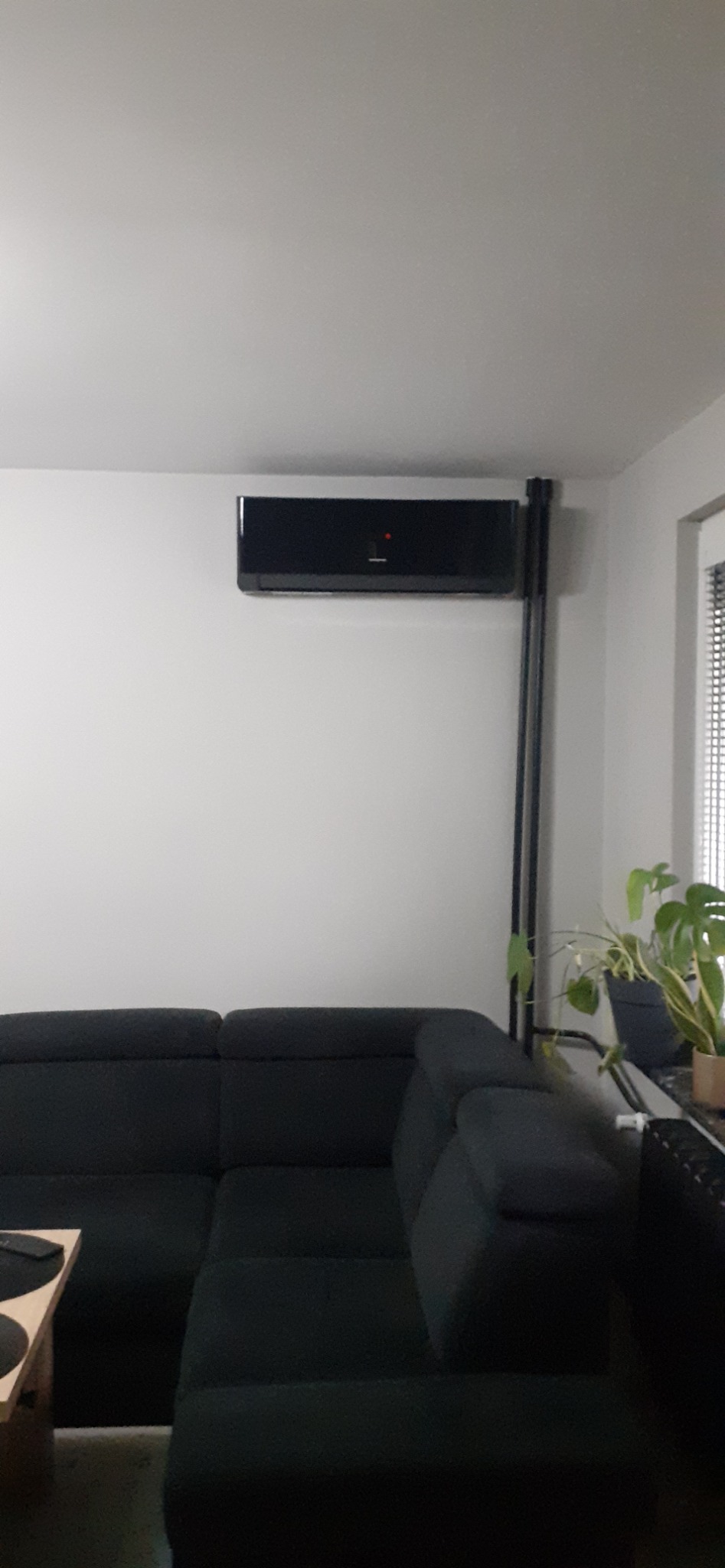Czarny klimatyzator ścienny w nowoczesnym salonie z ciemną sofą. Widoczna roślina doniczkowa na parapecie i minimalistyczny design wnętrza.