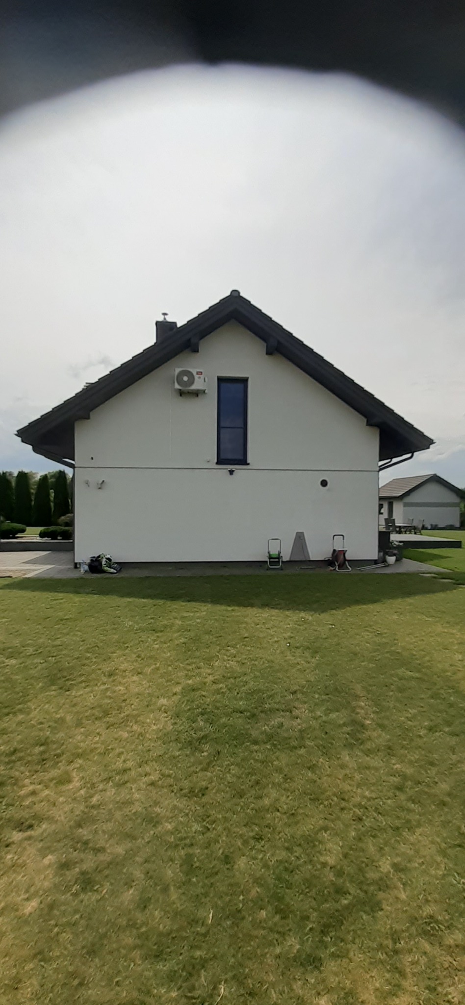 Biały dom z ciemnym dachem i zamontowaną klimatyzacją na ścianie. Widok z trawnika, w tle budynek gospodarczy i drzewa.