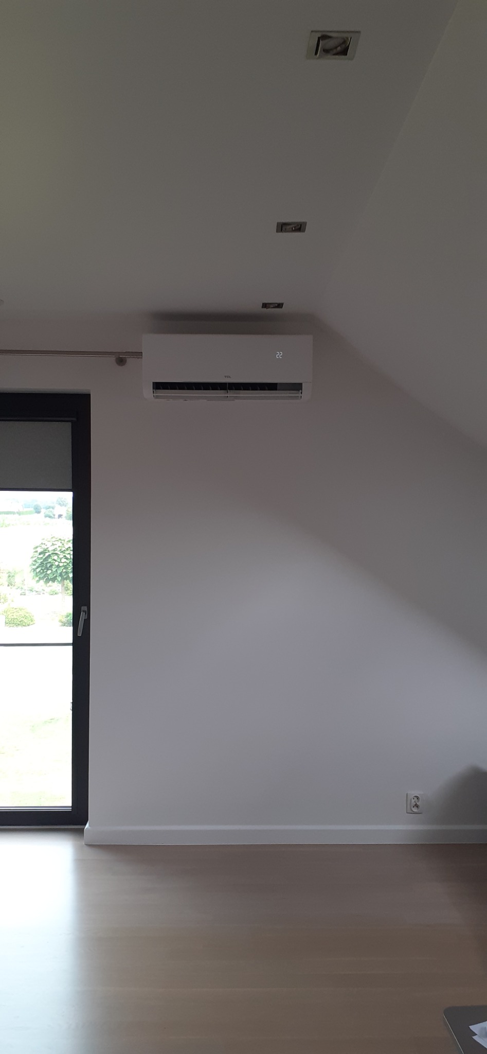 Biały klimatyzator na ścianie w jasnym pokoju z drewnianą podłogą. Widoczne okno z roletą i oświetlenie sufitowe.