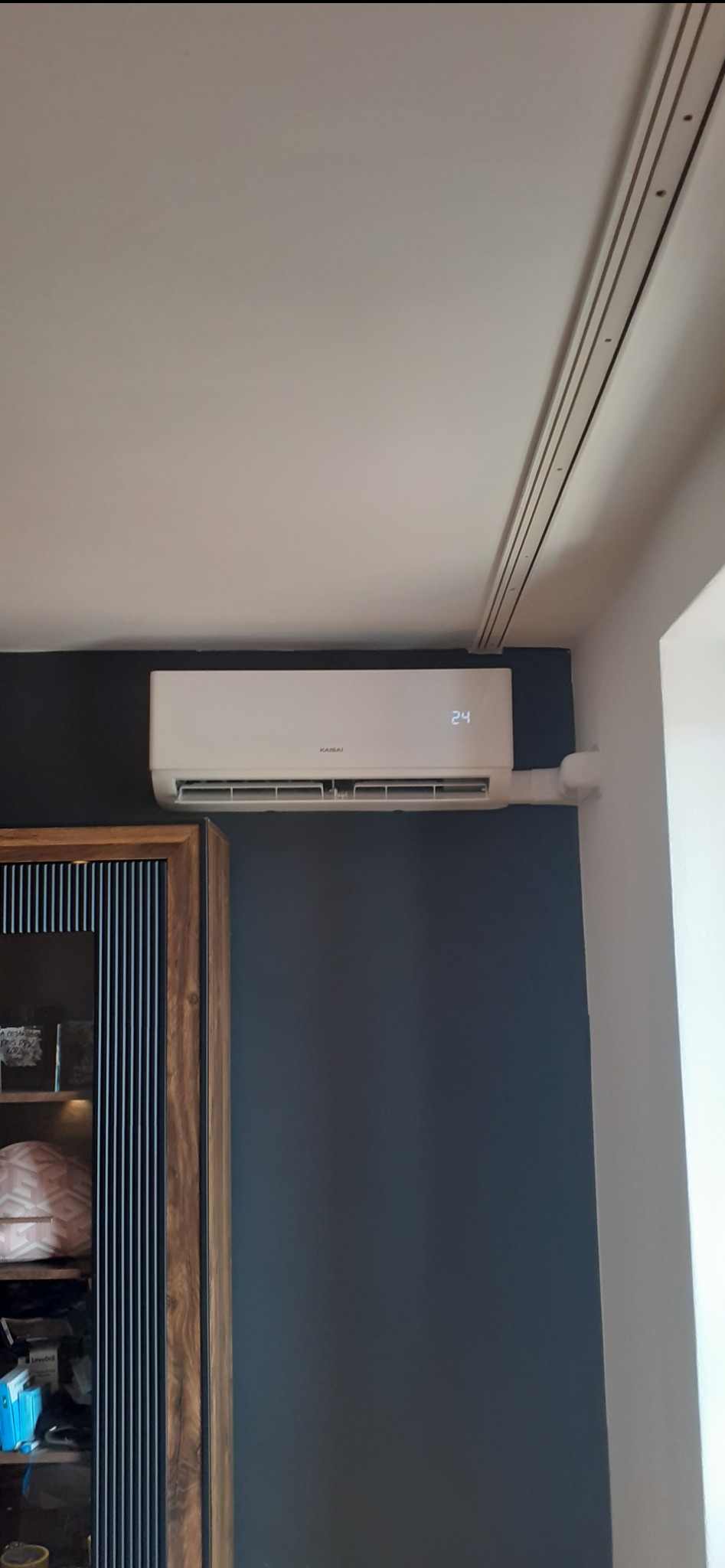 Biały klimatyzator Kaisai zamontowany na ciemnej ścianie w pokoju, widoczne rury instalacyjne i fragment mebla. Na wyświetlaczu urządzenia widoczna temperatura 24 stopnie.