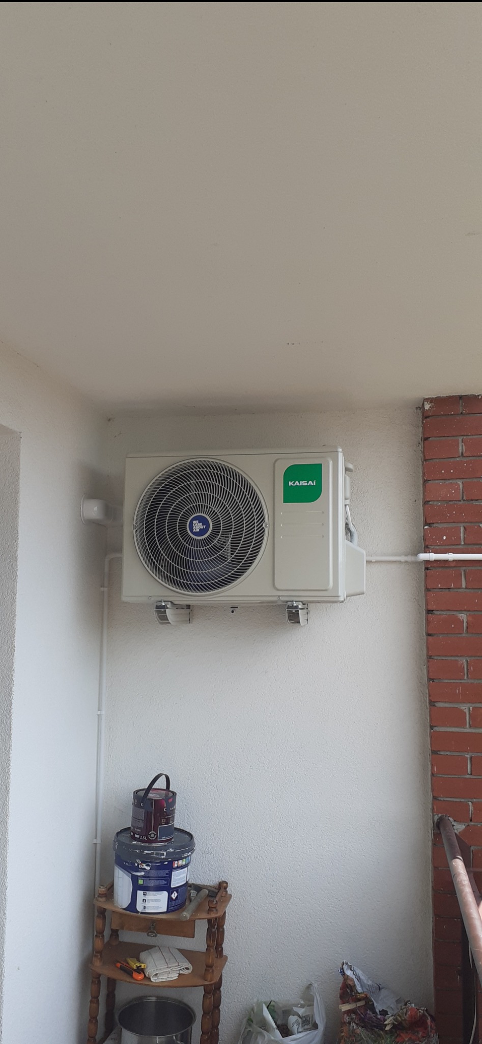 Zamontowany klimatyzator Kaisai na białej ścianie balkonu, z widocznymi rurami instalacyjnymi i stolikiem z farbami. Widok z dołu, jasne oświetlenie.