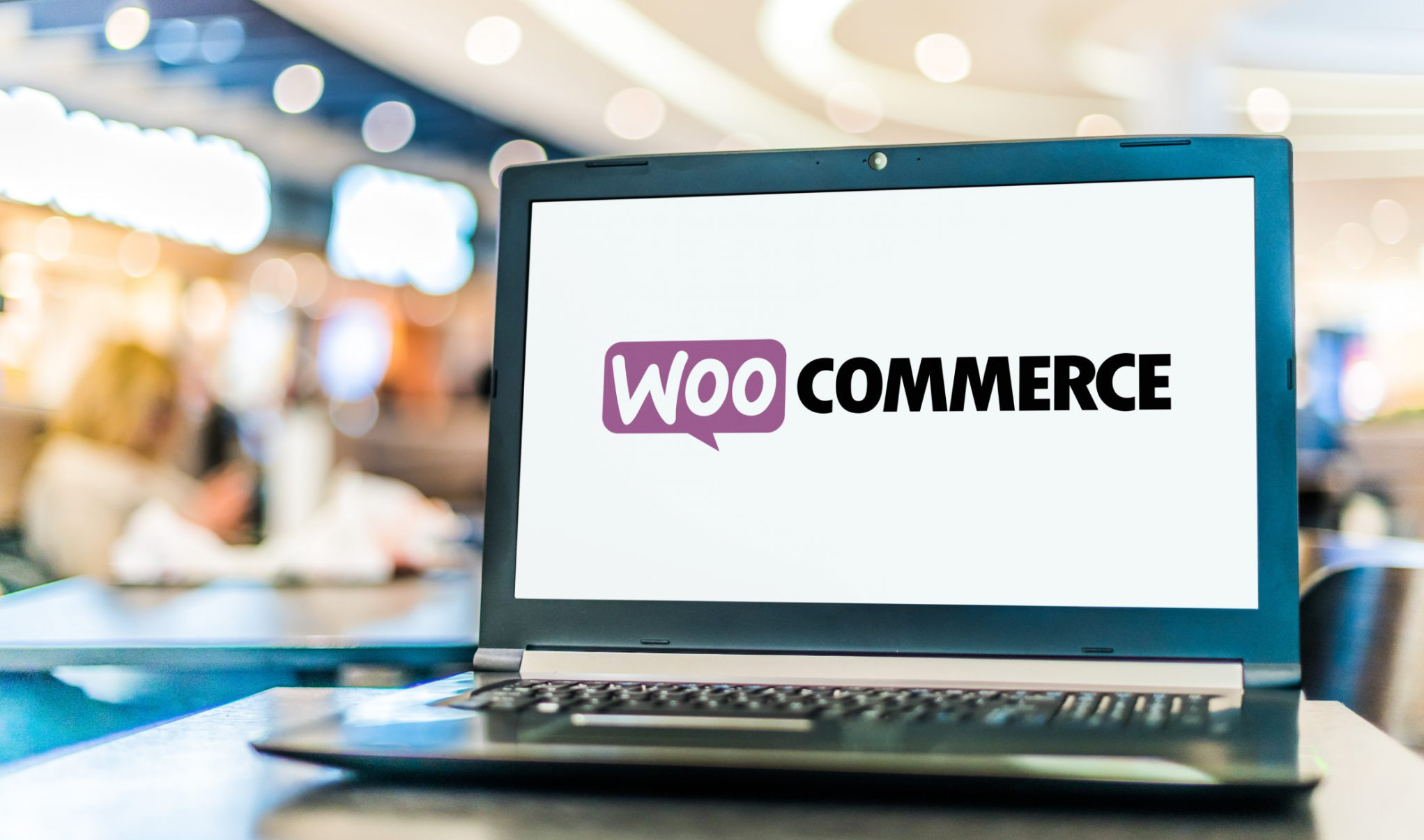 Laptop z logo WooCommerce na ekranie, ustawiony na stole w kawiarni. Widoczne rozmyte tło z ludźmi. Prezentacja platformy e-commerce.