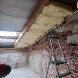 S-Bud - Poddasze w trakcie prac: widoczna izolacja z wełny mineralnej między krokwiami, drabina oparta o ceglaną ścianę. Częściowo otynkowana ściana z instalacją elektryczną.
