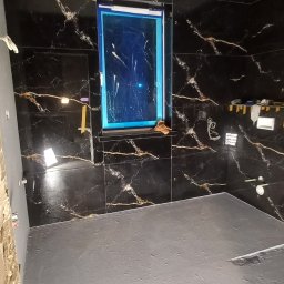 S-Bud - Łazienka w trakcie prac: czarne płytki imitujące marmur na ścianach, zabezpieczone okno folią, wylewka na podłodze. Widoczne narzędzia i niedokończone ściany.