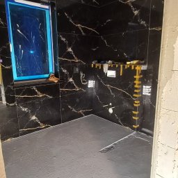 S-Bud - Ciemne płytki imitujące marmur na ścianach łazienki w trakcie prac. Podłoga pokryta szarą wylewką. Okno zabezpieczone folią. Widoczne taśmy zabezpieczające.