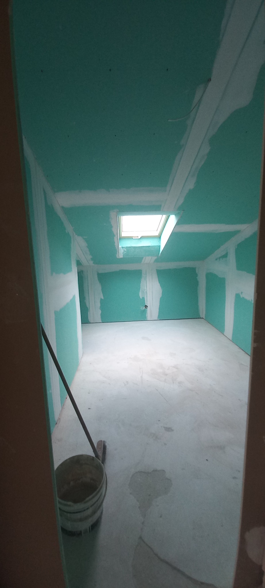 Pokój w trakcie wykańczania: zielone płyty gipsowo-kartonowe na ścianach i suficie, widoczne łączenia, okno dachowe. Na podłodze wiadro i narzędzia.