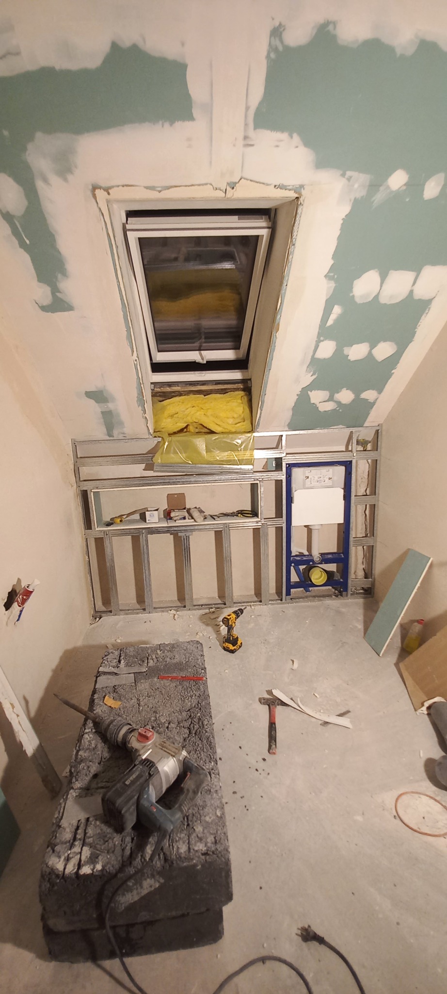 Poddasze w trakcie adaptacji: okno dachowe, metalowy stelaż pod zabudowę, częściowo zagruntowane ściany, narzędzia pracy na podłodze. Widok z dołu.