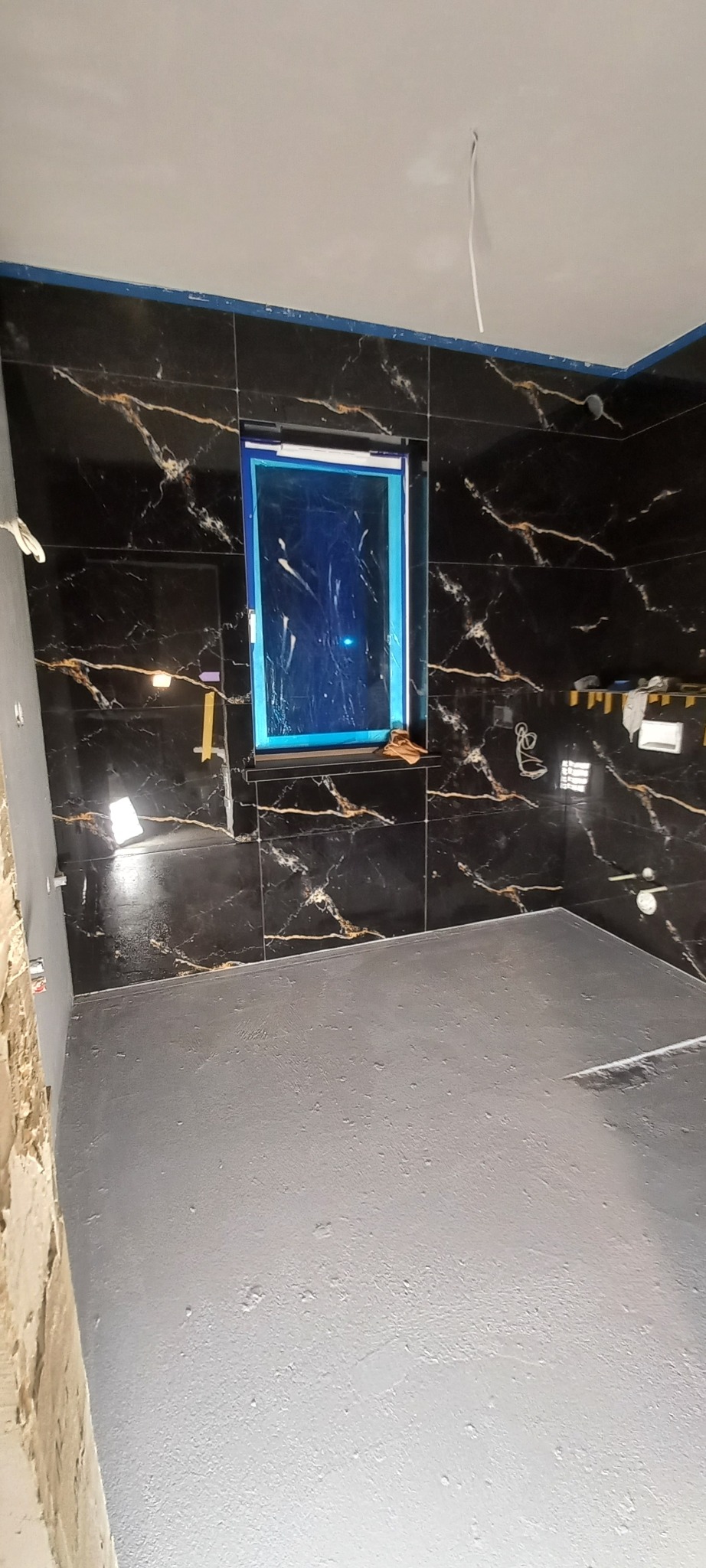 Łazienka w trakcie prac: czarne płytki imitujące marmur na ścianach, zabezpieczone okno folią, wylewka na podłodze. Widoczne narzędzia i niedokończone ściany.