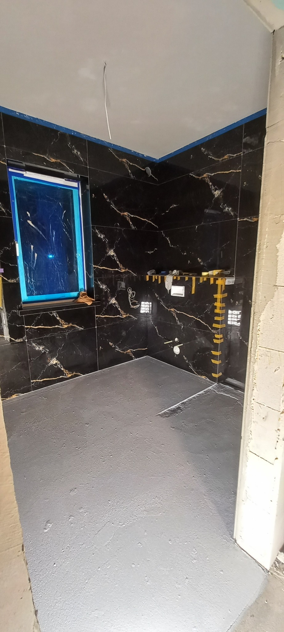 Ciemne płytki imitujące marmur na ścianach łazienki w trakcie prac. Podłoga pokryta szarą wylewką. Okno zabezpieczone folią. Widoczne taśmy zabezpieczające.