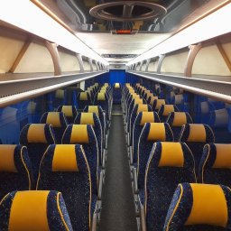 Luks Bus - Przewozy Pasażerskie Łukasz Muszyński - Wnętrze autobusu z rzędami niebieskich foteli z żółtymi zagłówkami, oświetlone jasnym światłem. Widok z perspektywy pasażera, symetryczny układ siedzeń.