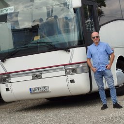 Luks Bus - Przewozy Pasażerskie Łukasz Muszyński - Biały autokar Van Hool z polską rejestracją SR-7635L, na tle szarego żwiru, z mężczyzną w niebieskiej koszuli i okularach przeciwsłonecznych, opierającym się o pojazd.