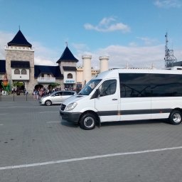 Luks Bus - Przewozy Pasażerskie Łukasz Muszyński - Biały bus na parkingu przed parkiem rozrywki EnergyLandia. Pojazd z przyciemnianymi szybami, widoczny z boku, na pierwszym planie. W tle budynki parku i spacerujący ludzie.