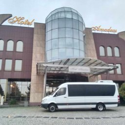 Luks Bus - Przewozy Pasażerskie Łukasz Muszyński - Biały bus z przyciemnianymi szybami zaparkowany przed hotelem Amadeus z przeszkloną kopułą. Transport gości hotelowych.