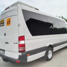 Luks Bus - Przewozy Pasażerskie Łukasz Muszyński - Biały bus Mercedes Sprinter CDI 515 z przyciemnianymi szybami i naklejką z symbolem dzieci, zaparkowany na kostce brukowej. Widok z tyłu i boku.