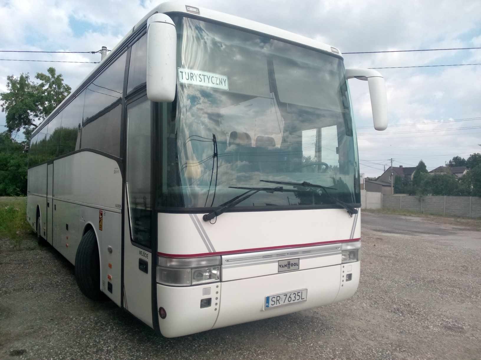 Biały autokar turystyczny marki Van Hool z napisem 'Turystyczny' na szybie, zaparkowany na żwirowym placu, z widocznym numerem rejestracyjnym SR 7635L.