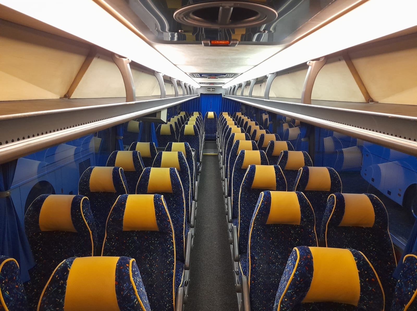 Wnętrze autobusu z rzędami niebieskich foteli z żółtymi zagłówkami, oświetlone jasnym światłem. Widok z perspektywy pasażera, symetryczny układ siedzeń.